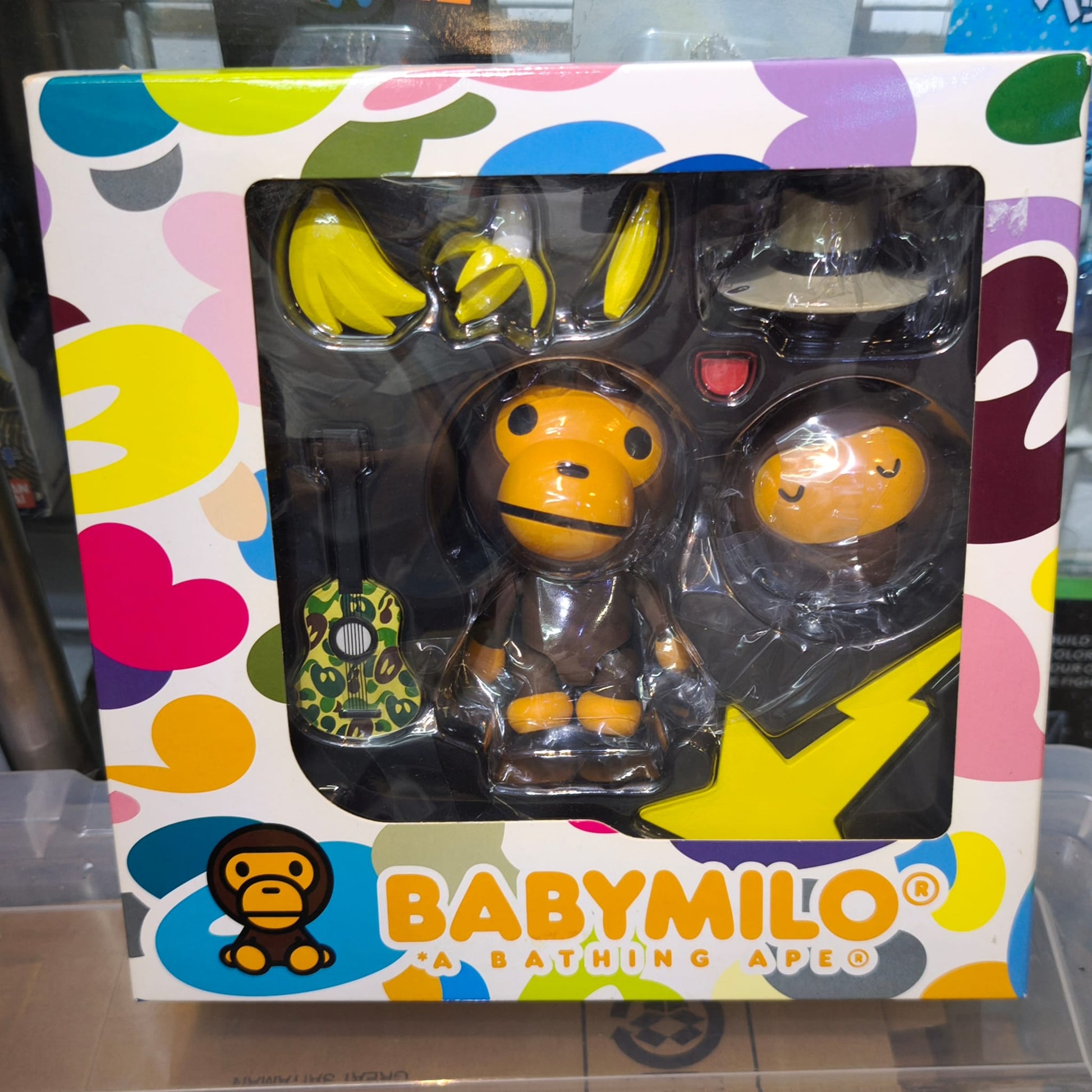 The back of the colorful green camo packaging for the Kaiyodo Revoltech Yamaguchi No. 115 Baby Milo figure, showcasing accessories including the guitar, magnetic hat, banana variations, interchangeable face, and the magnetic foot design.